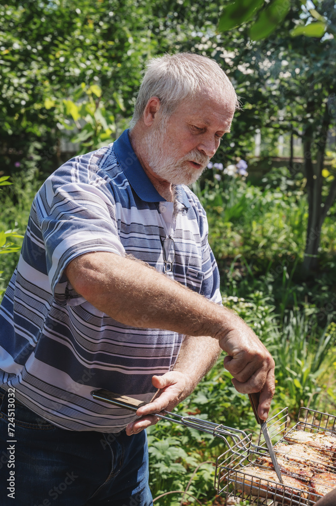 Fototapeta premium An elderly gray-haired man with a beard is cooking meat on a grate and open fire in the garden. Weekend vacation, cooking