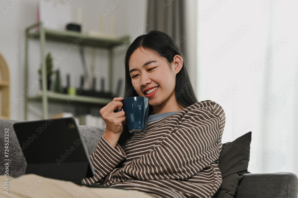 Carefree young woman drinking hot tea and using digital tablet on couch at home Carefree young woman drinking hot tea and using digital tablet on couch at home