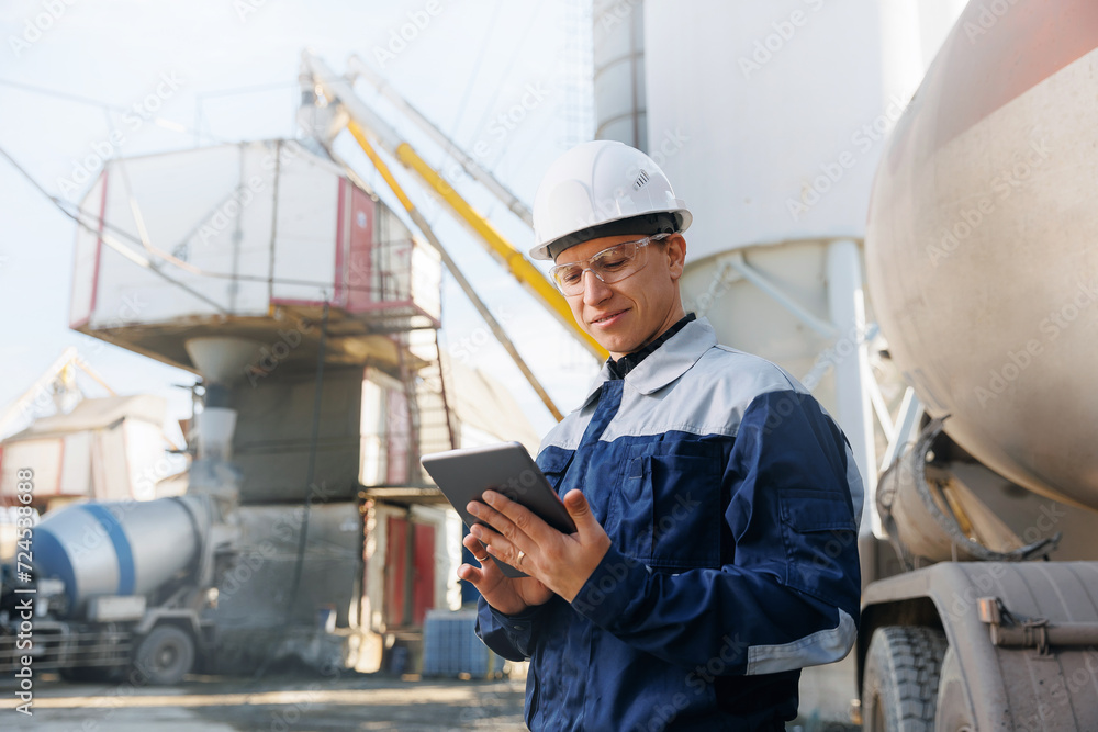 Engineer man in uniform with tablet computer controls loading of cement to mixer truck on concrete factory. Concept automated logistics on industry with online internet