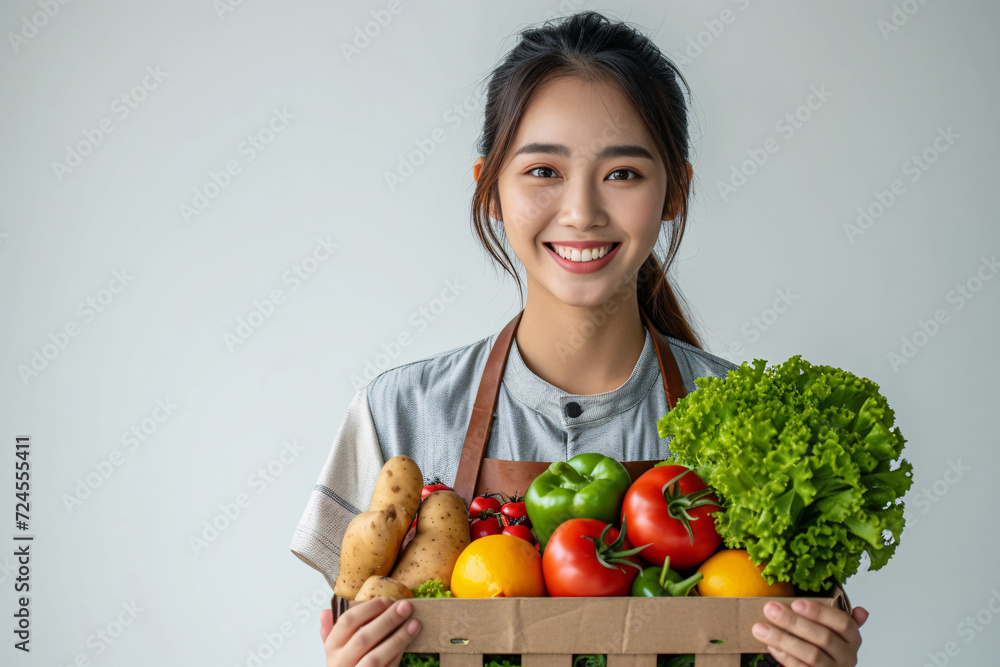 Smiling young woman in apron holding box filled with fresh vegetables and fruits on light Smiling young woman in apron holding box filled with fresh vegetables and fruits on light