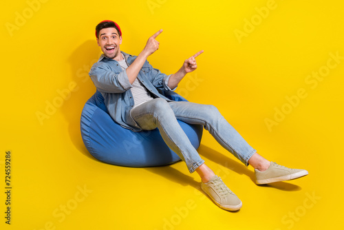 Full length photo of excited funky guy wear denim jacket relaxing bean bag showing two fingers empty space isolated yellow color background