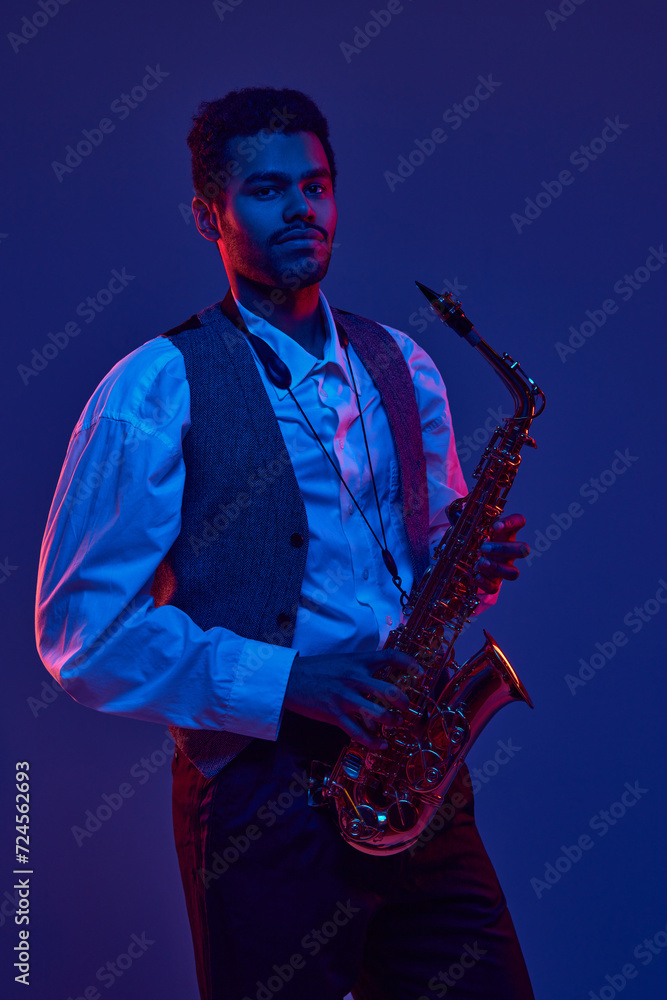 Naklejka premium Portrait of young man, solo performer, instrumental musician holds saxophone and looking at camera against dark background in neon light. Concept of jazz, blues, classy instrumental music, concert. Ad