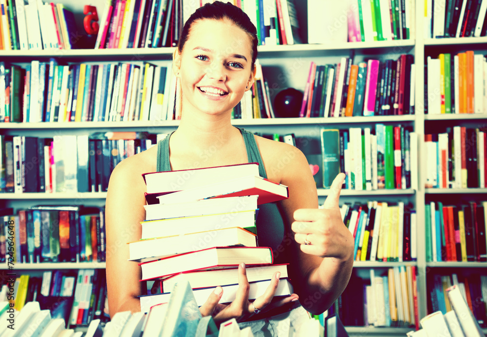 Girl holding stack of books shows thump up in a bookstore Girl holding stack of books shows thump up in a bookstore