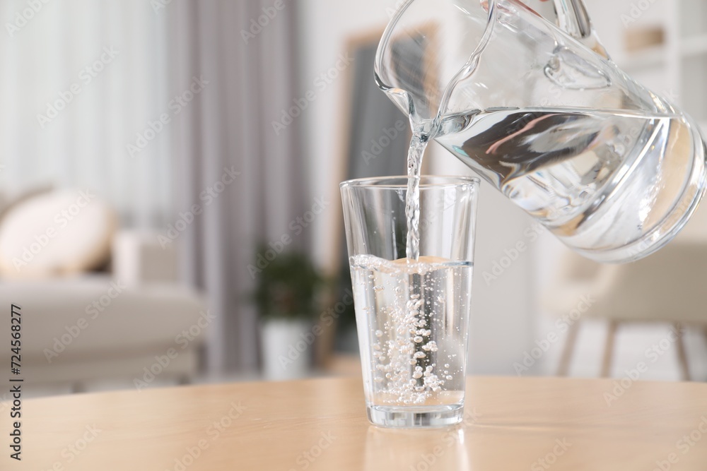 Pouring fresh water from jug into glass at table against blurred background, closeup. Space for text Pouring fresh water from jug into glass at table against blurred background, closeup. Space for text
