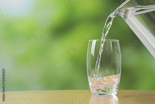 Pouring fresh water from jug into glass at wooden table against blurred green background, closeup. Space for text