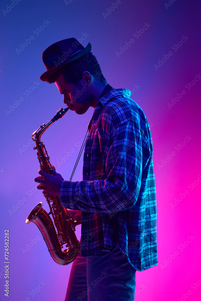 Naklejka premium Portrait of talented solo saxophonist playing piece against pink-blue gradient background in neon light. Concept of jazz, blues, classy instrumental music, festivals and concerts. Ad