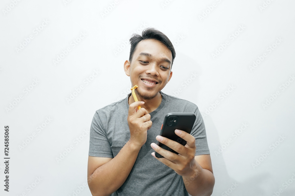 Happy laughing Asian man shaving his beard and holding smartphone Happy laughing Asian man shaving his beard and holding smartphone
