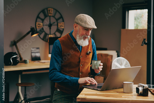 Hipster assis souriant quinquagénaire senior élégant et stylé qui travaille sur un ordinateur dans un atelier créatif vintage et qui porte un gilet et une casquette et qui bois un café photo