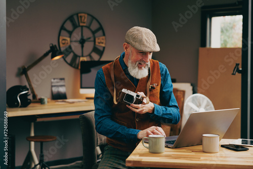 Homme photographe assis souriant quinquagénaire senior hipster élégant et stylé qui travaille sur un ordinateur dans un atelier créatif vintage et qui tient un appareil photo en buvant un café photo