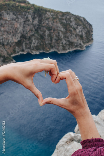 female hands folded in the shape of a heart against the background of rocks and the blue Mediterranean sea