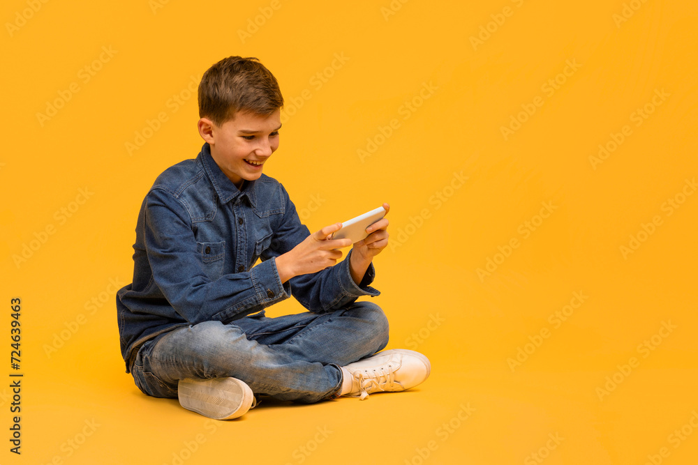Cheerful teen boy playing games on smartphone while sitting on yellow background Cheerful teen boy playing games on smartphone while sitting on yellow background