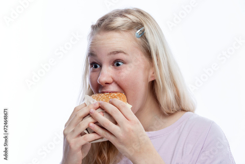 Smiling hungry teenage girl takes a bite of hamburger. Delicious and popular fast food. A cute blonde with freckles in a pink jacket. White background.