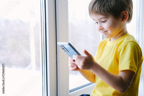 Little toddler boy is holding the smartphone at home. He is wearing bright yellow t-shirt. Playing video games like a social problem photo