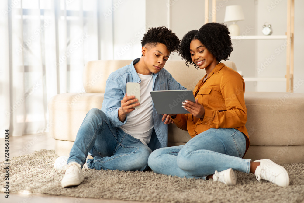 Casually dressed in denim, a young couple sits on a plush rug, engrossed in their devices Casually dressed in denim, a young couple sits on a plush rug, engrossed in their devices