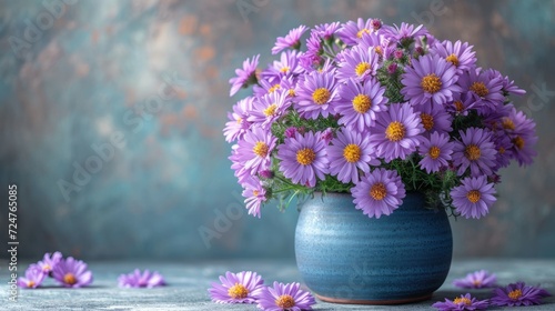  a blue vase filled with purple flowers sitting on top of a wooden table next to other purple and yellow flowers on a gray and blue tablecloth covered tablecloth.