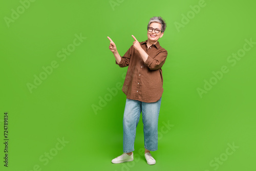 Full length photo of positive woman dressed oversize shirt jeans indicating at discount empty space isolated on green color background © deagreez