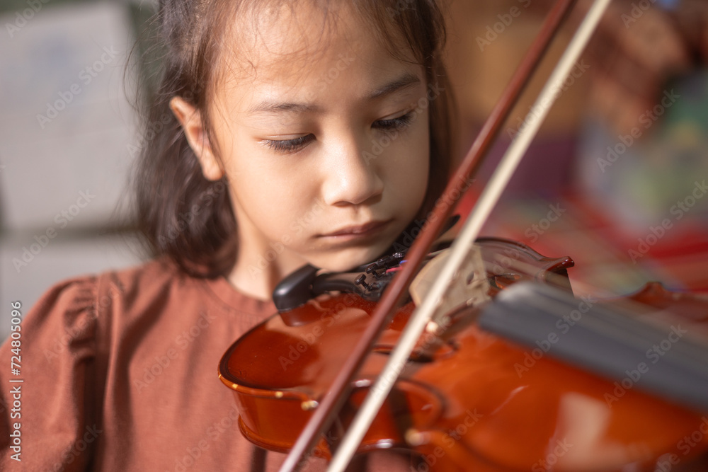 Close up face of cute Asian kid girl playing beautiful violin showing her face with determination for musical practice at home. Her small hands move bow across the wooden strings for beautiful song. Close up face of cute Asian kid girl playing beautiful violin showing her face with determination for musical practice at home. Her small hands move bow across the wooden strings for beautiful song.