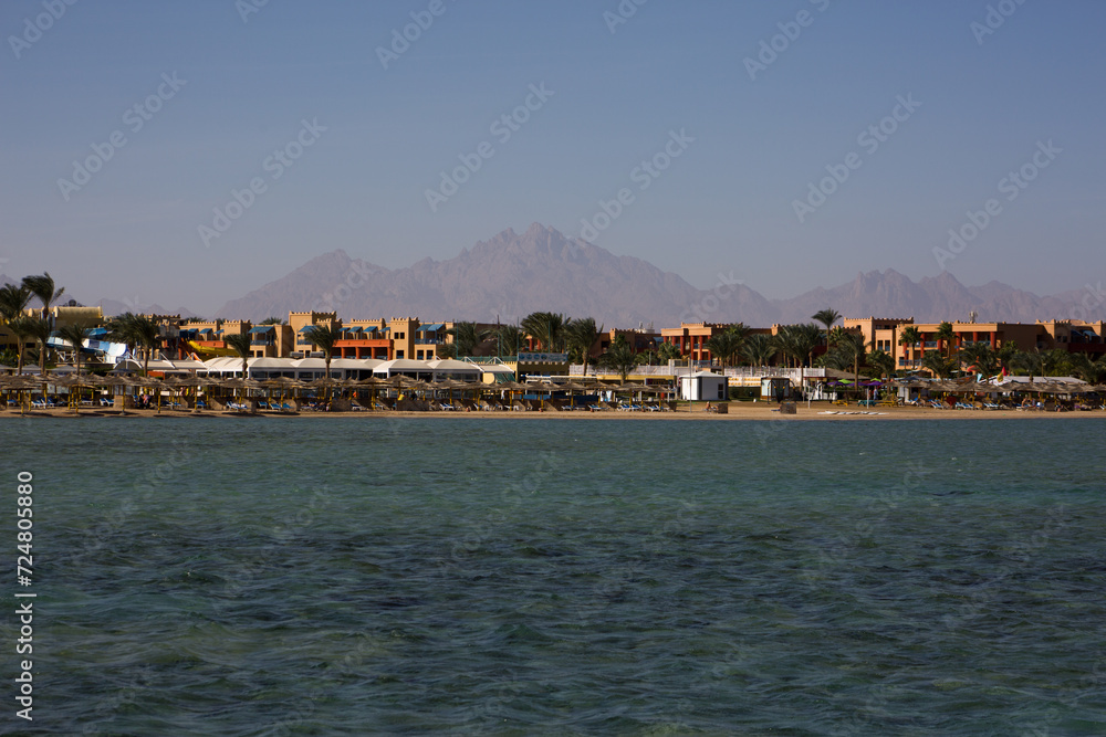 Beach in Egypt with places to relax near the hotel with palm trees. Red sea and sandy beach on a sunny day with a view of equipped seating areas under an umbrella
