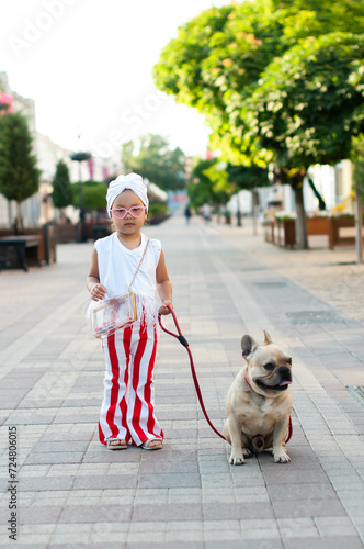 little pretty girl stylishly dressed with a white turban on her head and red and white striped pants walking her French bulldog dog on a city street.. photo