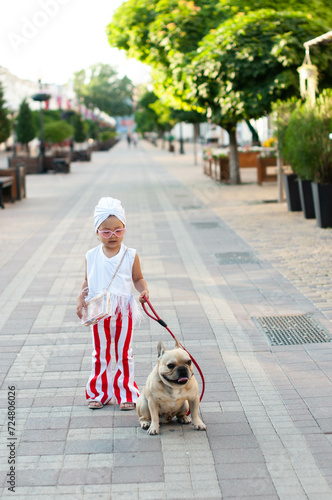 little pretty girl stylishly dressed with a white turban on her head and red and white striped pants walking her French bulldog dog on a city street.. photo