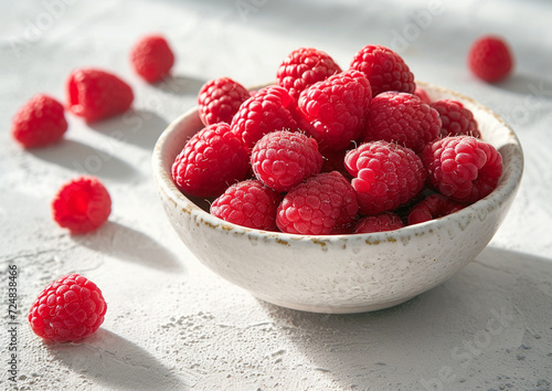 raspberry in a white ceramic bowl, with a few raspberries scattered around the bowl, on a white background