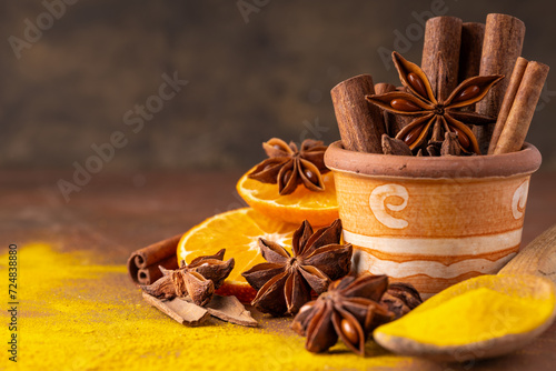 In the foreground various spices and orange slices. In a terracotta jar some cinnamon sticks and a star anise star. Still life