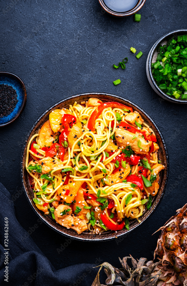 Stir fryed egg noodles with chicken, pineapple, paprika, green onion, soy sauce and sesame seeds in bowl. Asian cuisine dish. Black table background, top view Stir fryed egg noodles with chicken, pineapple, paprika, green onion, soy sauce and sesame seeds in bowl. Asian cuisine dish. Black table background, top view