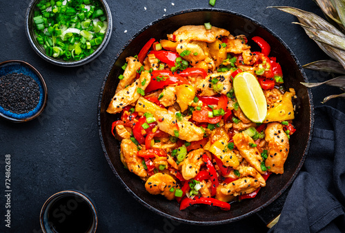 Stir fry chicken slices with pineapple, paprika, green onion, soy sauce and sesame seeds in frying pan. Asian cuisine dish. Black table background, top view