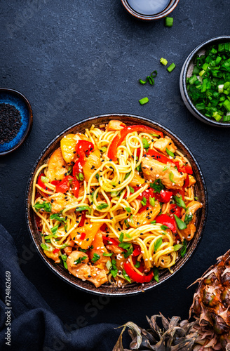 Stir fryed egg noodles with chicken, pineapple, paprika, green onion, soy sauce and sesame seeds in bowl. Asian cuisine dish. Black table background, top view