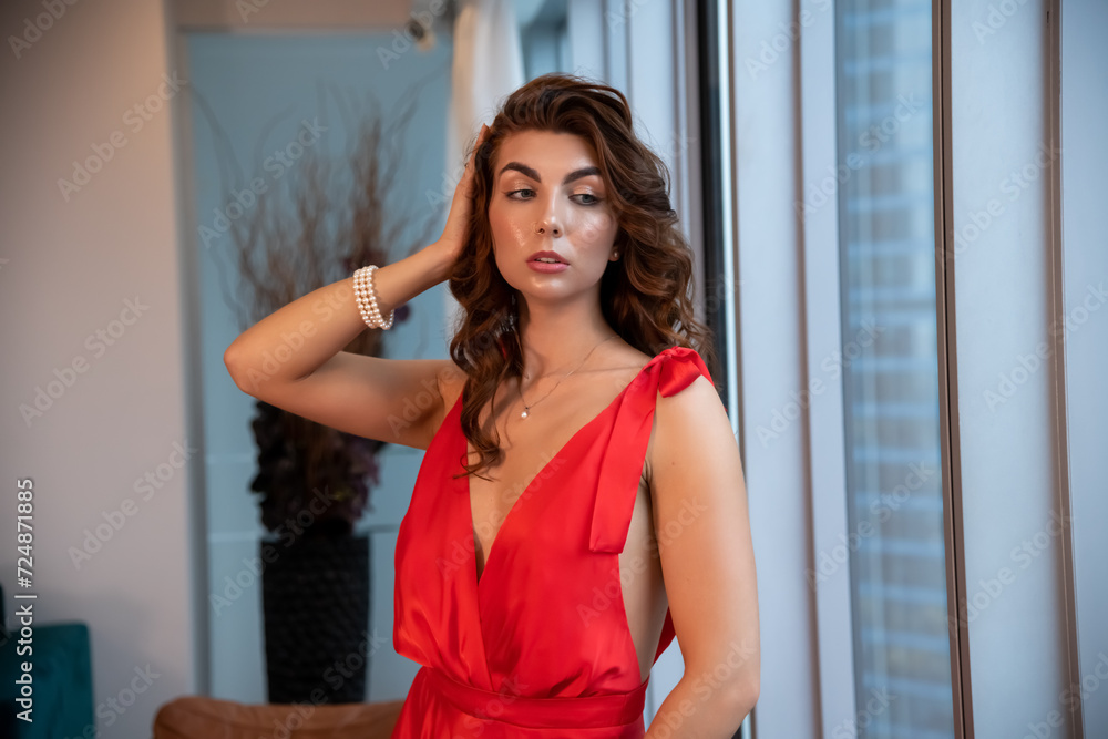 Waist up portrait of beautiful caucasian brunette woman in long red dress standing by apartment's window. Soft focus. Beauty and fashion theme.