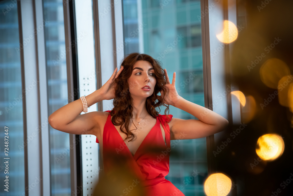 Waist up portrait of beautiful caucasian brunette woman in long red dress standing by apartment's window. Blurred Christmas tree with orange Christmas lights in the foreground. Winter holidays theme.