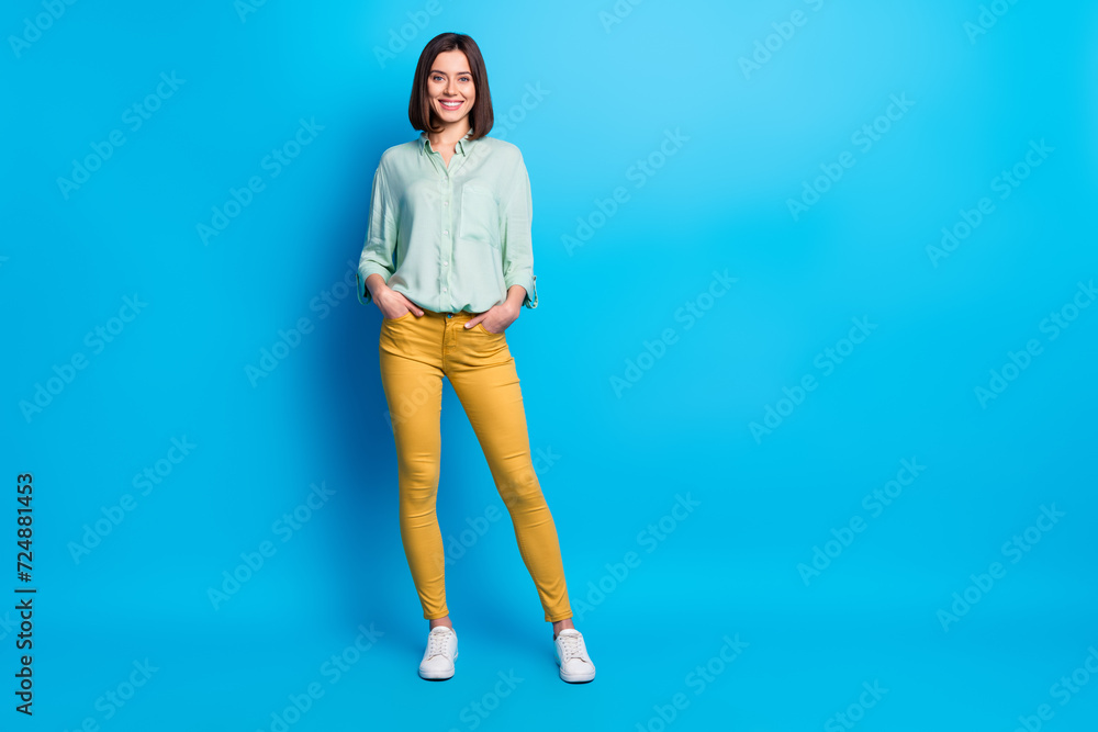 Full size photo of positive cool pretty girl with stylish hair wear teal shirt standing arms in pockets isolated on blue color background Full size photo of positive cool pretty girl with stylish hair wear teal shirt standing arms in pockets isolated on blue color background