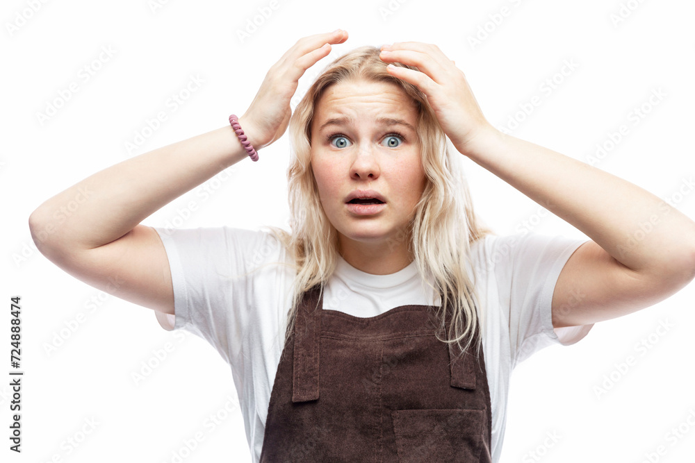 The teenage girl raised her hands up in shock. Cute blonde girl in a white T-shirt and brown overalls. Horror and surprise. White background.