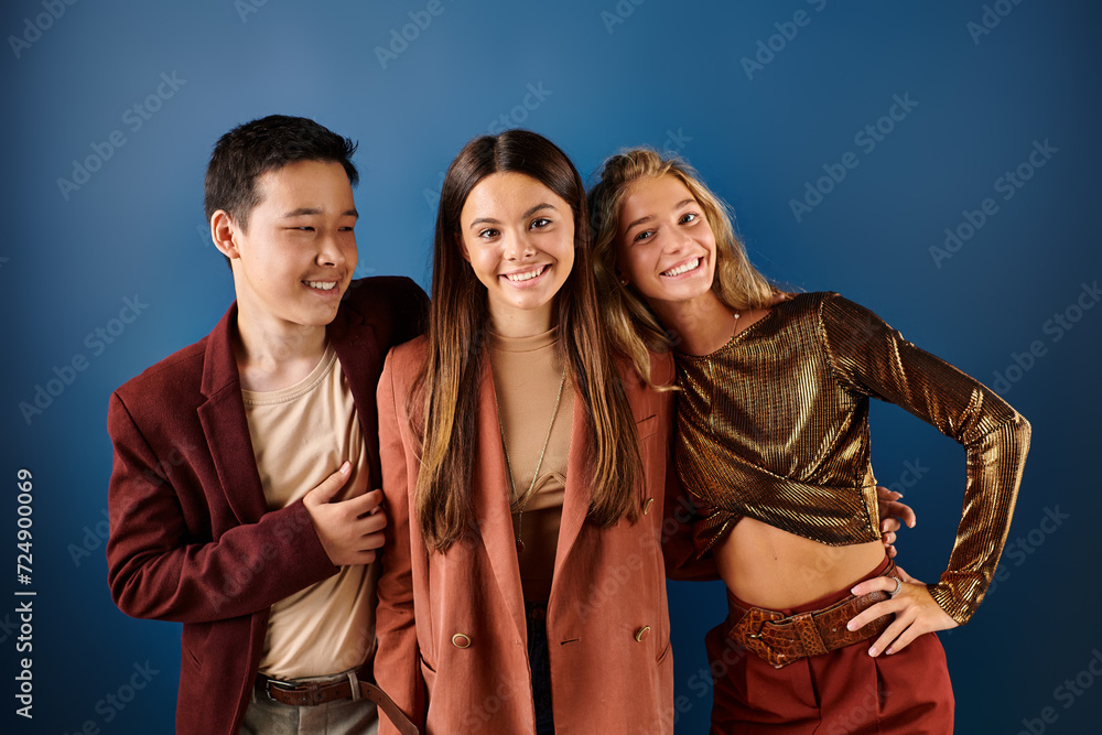 jolly multiracial adolescent friends in vivid attires posing on blue backdrop and smiling joyfully jolly multiracial adolescent friends in vivid attires posing on blue backdrop and smiling joyfully