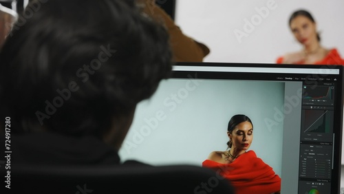 Backstage of model and professional team in the studio. Model posing on set, male editor working on computer looking at photos on monitor.