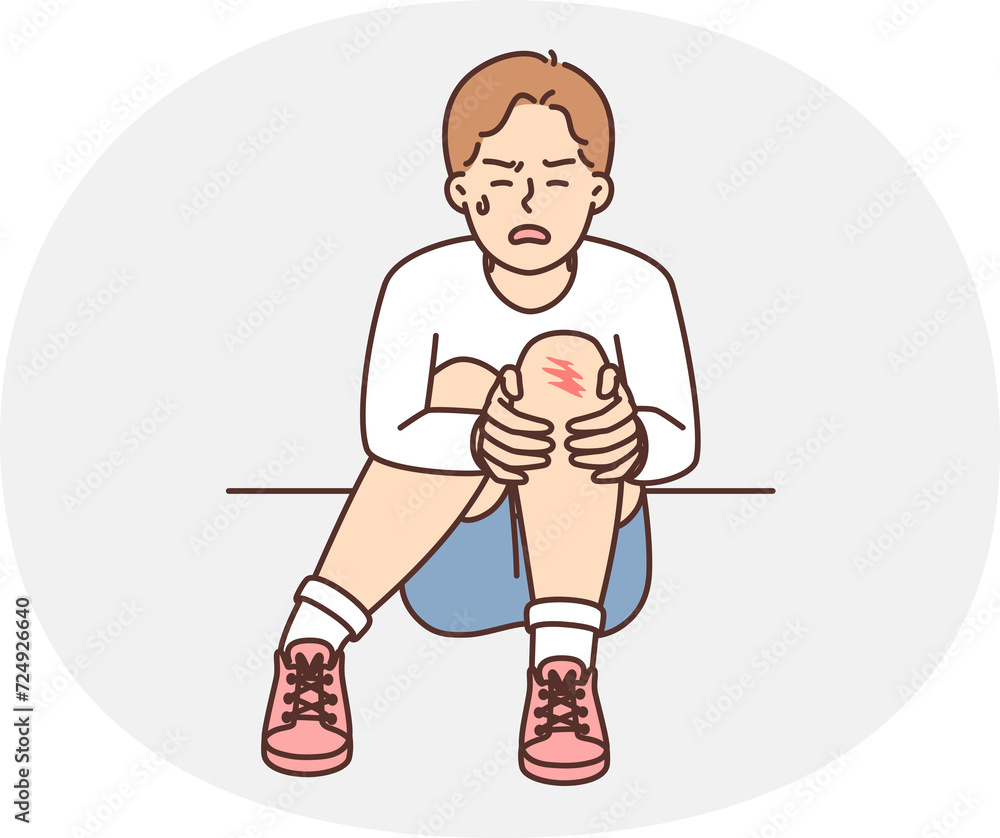 Crying teenager boy sits on ground and holds on to injury on knee after fall during walk. Unfortunate child was injured during sports activities or active games with friends Crying teenager boy sits on ground and holds on to injury on knee after fall during walk. Unfortunate child was injured during sports activities or active games with friends