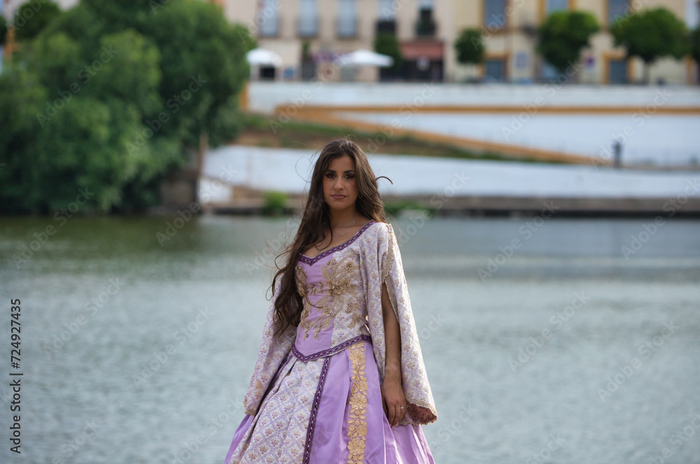 Beautiful latin woman with long curly hair wearing a 15th century dress is standing by the banks of the river guadalquivir in seville in spain. In the background you can see the other side of the city