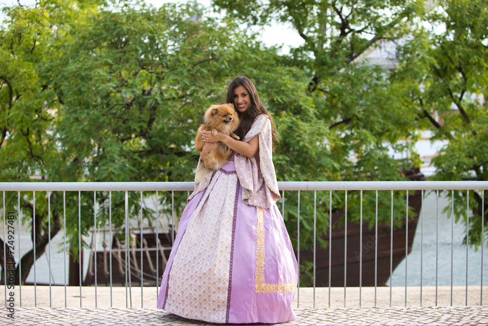 Fototapeta premium Beautiful Latin woman with long curly hair wearing a 15th century dress is holding a pomenarian dog in her arms. The woman is happy with her pet. Dog and pet day concept.