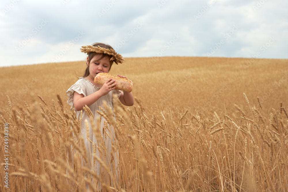 A little girl stands among the wheat ears with bread in her hands. She takes a bite from the bread product. Copy space.