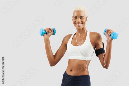 smiling woman doing fitness workout, lifts dumbbells while listening music through earphones of mobile phone on arm band. Latin American female athlete. Sportswoman do training, isolated on background © amedeoemaja