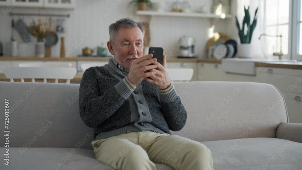 Laughing senior man talking chatting video call on smartphone sitting on couch at home looking at screen. Old pensioner enjoys online conversation with relatives, friends. Emotionally speaking phone. Laughing senior man talking chatting video call on smartphone sitting on couch at home looking at screen. Old pensioner enjoys online conversation with relatives, friends. Emotionally speaking phone.