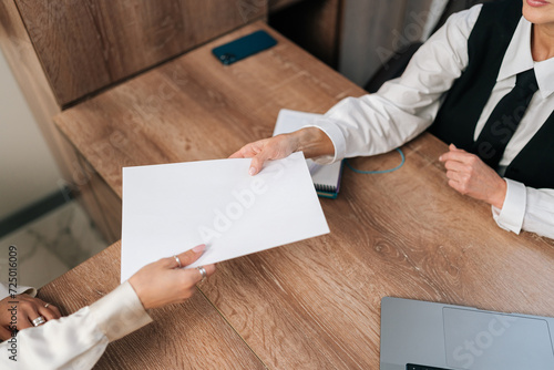 Top view of middle-aged businesswoman manager passing papers documents to unrecognizable female colleague, sitting at table in office. Two business partners having meeting in conference room.