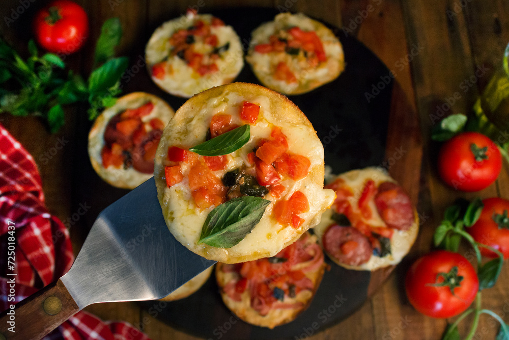 Small cheese and pepperoni pizzas with tomatoes, with a spatula holding one of the pizzas, on a wooden board and in the background a red and white checkered cloth, blurred tomatoes and seasonings