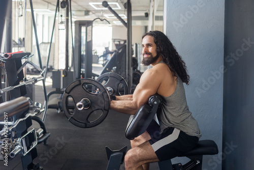 Healthy lifestyle and body care. Profile portrait of attractive bodybuilder with long curly hair having workout in gym lifting barbell, doing exercises for biceps and triceps. Indoor shot.