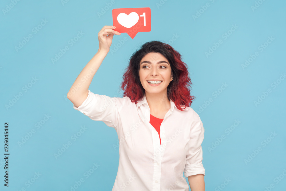 Portrait of joyful cheerful woman with fancy red hair holding blogger heart above head, communicating online, wearing white shirt. Indoor studio shot isolated on blue background. Portrait of joyful cheerful woman with fancy red hair holding blogger heart above head, communicating online, wearing white shirt. Indoor studio shot isolated on blue background.