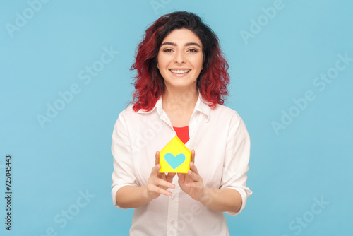 Portrait of attractive cheerful woman with fancy red hair holding paper house, mortgage, real estate agency, wearing white shirt. Indoor studio shot isolated on blue background. © khosrork