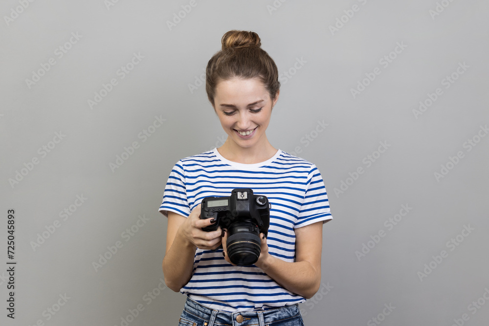 Portrait of satisfied delighted woman traveler or photographer wearing striped T-shirt holding and looking at photocamera, taking photo enjoying hobby. Indoor studio shot isolated on gray background. Portrait of satisfied delighted woman traveler or photographer wearing striped T-shirt holding and looking at photocamera, taking photo enjoying hobby. Indoor studio shot isolated on gray background.