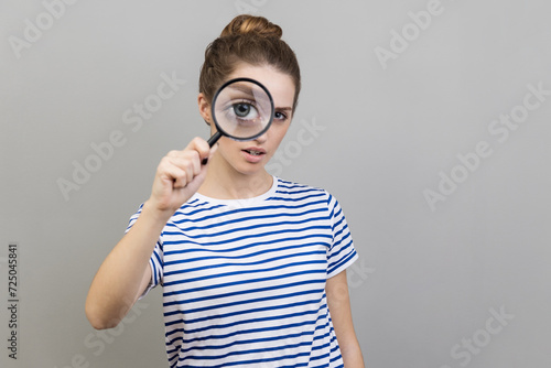 Portrait of woman wearing striped T-shirt holding magnifying glass and looking at camera with big zoom eye, serious face. Indoor studio shot isolated on gray background. photo