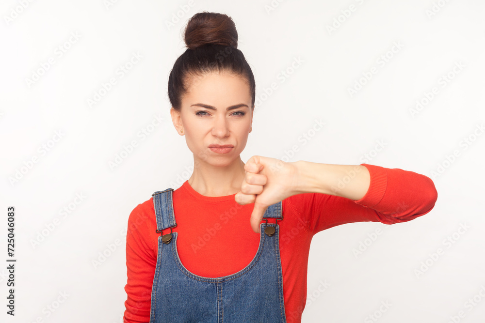 Portrait of sad disappointed woman with hair bun frowning face showing thumb down dislike gesture disapprove sign, wearing denim overalls. Indoor studio shot isolated on white background Portrait of sad disappointed woman with hair bun frowning face showing thumb down dislike gesture disapprove sign, wearing denim overalls. Indoor studio shot isolated on white background