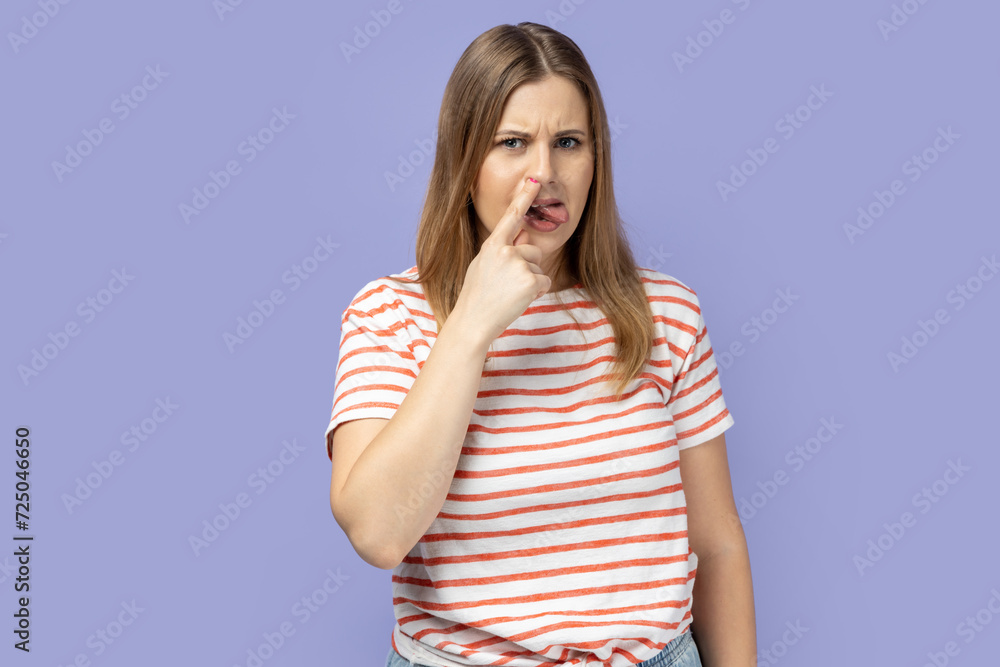 Portrait of blond woman wearing striped T-shirt putting finger into her nose and showing tongue, fooling around, bad habits, disrespectful behavior. Indoor studio shot isolated on purple background. Portrait of blond woman wearing striped T-shirt putting finger into her nose and showing tongue, fooling around, bad habits, disrespectful behavior. Indoor studio shot isolated on purple background.